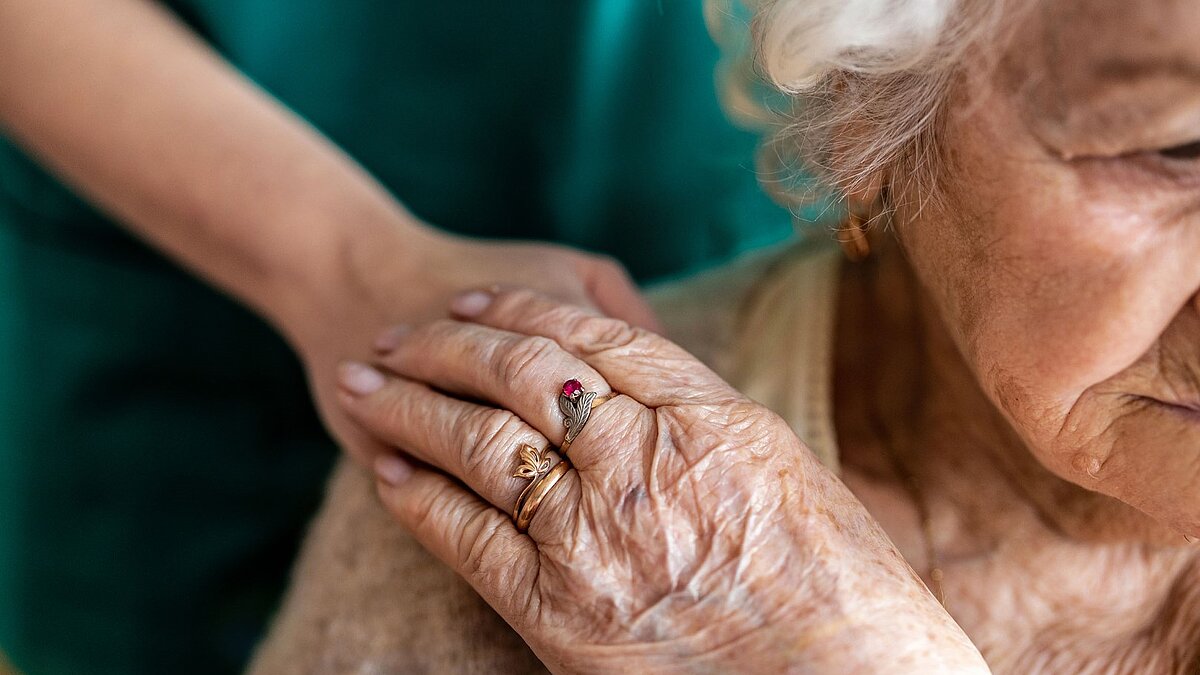 foto zeigt eine ältere frau die von einer nicht zu sehenden frau an der schulter berührt wird, die ältere frau legt ihre hand auf die der anderen person