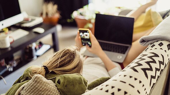 foto zeigt eine frau auf der couch mit smartphone und laptop